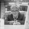 A black and white image of Prof. Cronin, smiling at the camera. He is wearing a grey suit and leaning over a lab bench towards the camera.