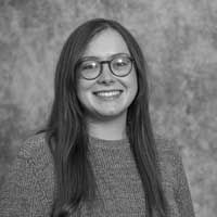A black and white image of Kanipe, smiling at the camera. She has long brown hair and is wearing a grey jumper.