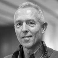 A black and white image of Meya, smiling at the camera. He has short white hair and is wearing a button-up shirt and zip-up fleece.