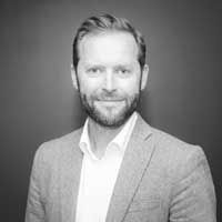 A black and white image of Wiklund, smiling at the camera. He has short hair and a well-groomed beard and is wearing a light-gray suit.