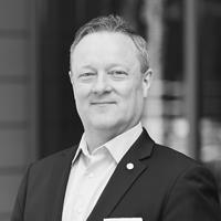 A black and white image of Baber, smiling at the camera. He has short, cropped gray hair and is wearing a white shirt with a dark-colored suit.