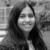 A black and white headshot of Varshney, smiling at the camera. She is an Indian woman with long, straight black hair and is wearing a dark-colored patterned blazer over a light-colored top.