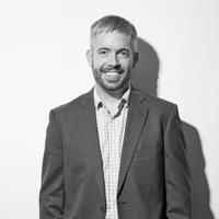 A black and white image of Cameron, smiling at the camera. He is a slim white man, wearing a light-colored check shirt and a gray suit. He has cropped gray hair and a short beard. 