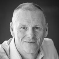 A black and white image of Brown, smiling at the camera. He has short gray-white hair and is wearing a white shirt.