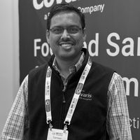 Black-and-white headshot of a smiling man wearing a vest and conference lanyard in front of a branded display.
