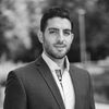 A black and white image of Khaled, smiling at the camera. He wears a white shirt and dark-gray suit, and has short black hair and stubble.