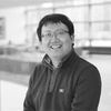 A black and white image of Cao, smiling at the camera. He has short black hair, wears glasses, and is wearing a dark-colored fleece jacket.