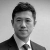 A black and white image of Shyu, smiling at the camera. He is wearing a dark colored suit, a white shirt, and a dark colored tie.