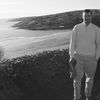 Man standing by coastal landscape with hills and shoreline in background.