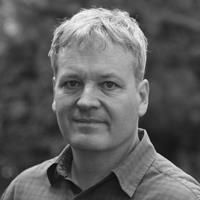 A black and white image of Hutterli, smiling at the camera. He has short white hair and is wearing a linen shirt.