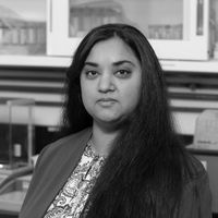 Headshot of a person with long dark hair wearing a blazer, seated indoors in an office setting.